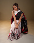 A model seated gracefully in a contemporary black and white crepe saree featuring bold red abstract brush strokes and a flowy silhouette.
