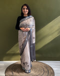 A model standing elegantly in a charcoal brown cotton saree featuring minimal black floral motifs and a structured striped border against a solid olive background.